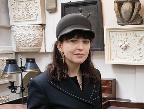 Nika Neelova, wearing a black jacket and grey hat, stands in the Drawing Office at Sir John Soane's Museum.