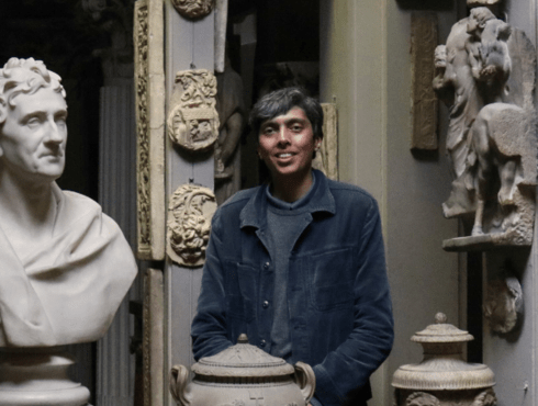Mohammed Qasim Ashfaq stands in the Dome area of Sir John Soane's Museum, next to a bust of Sir John Soane. He is wearing a blue shirt and smiling.