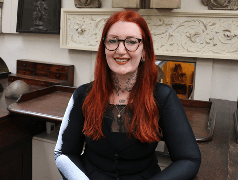 Lacey Law sits in Sir John Soane's Drawing Office, she is smiling at the camera. Lacey has red hair and is wearing glasses and a black top.